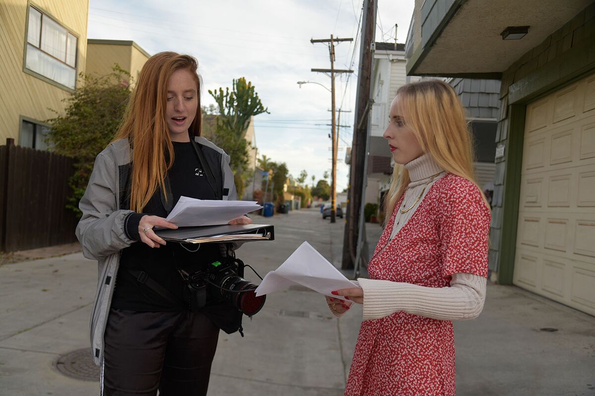 A director holding a camera and a script binder goes over pages with an actress in a red dress in an alley