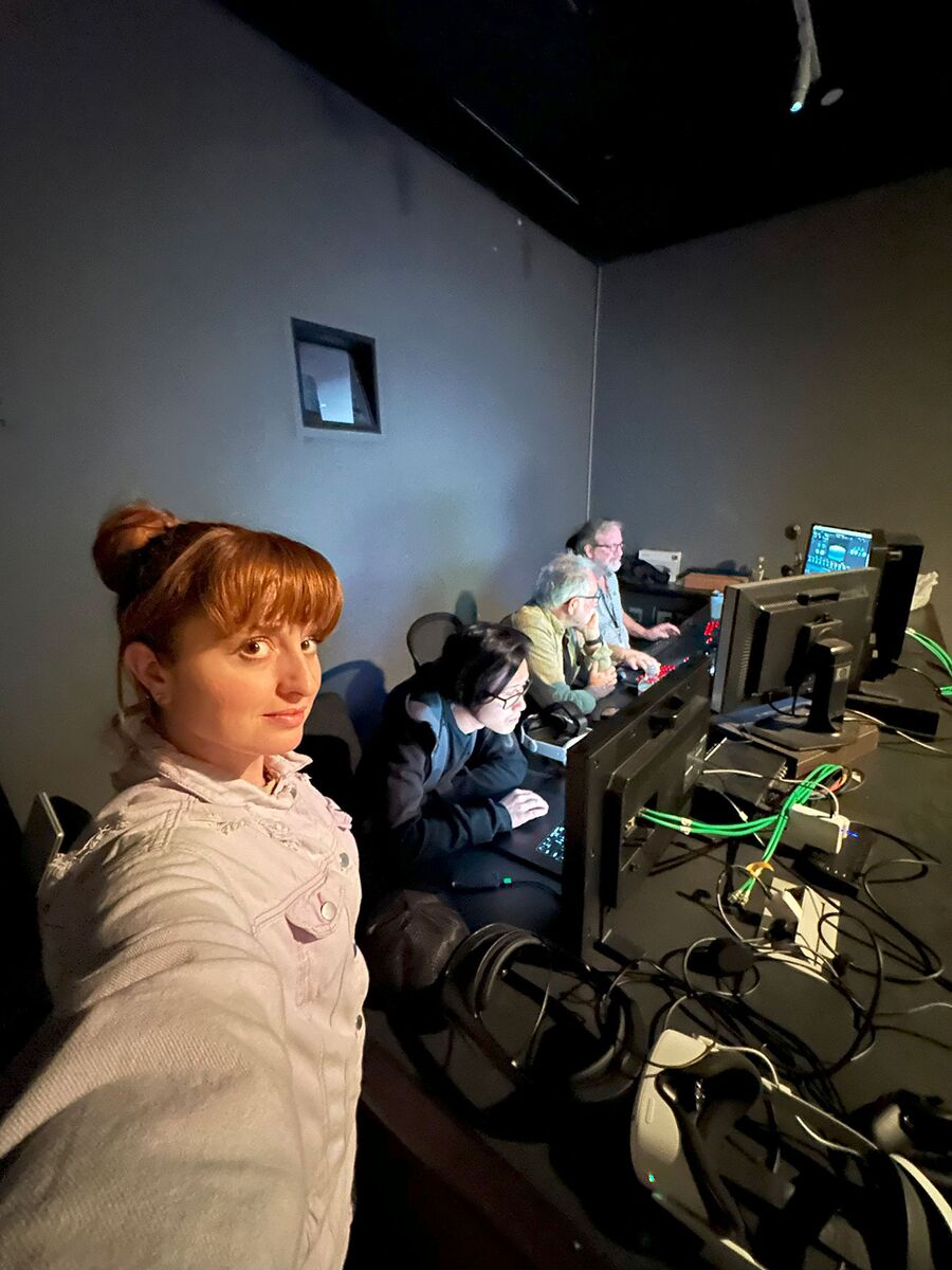 A crew member takes a selfie in a dark control room while three operators work at monitors behind her