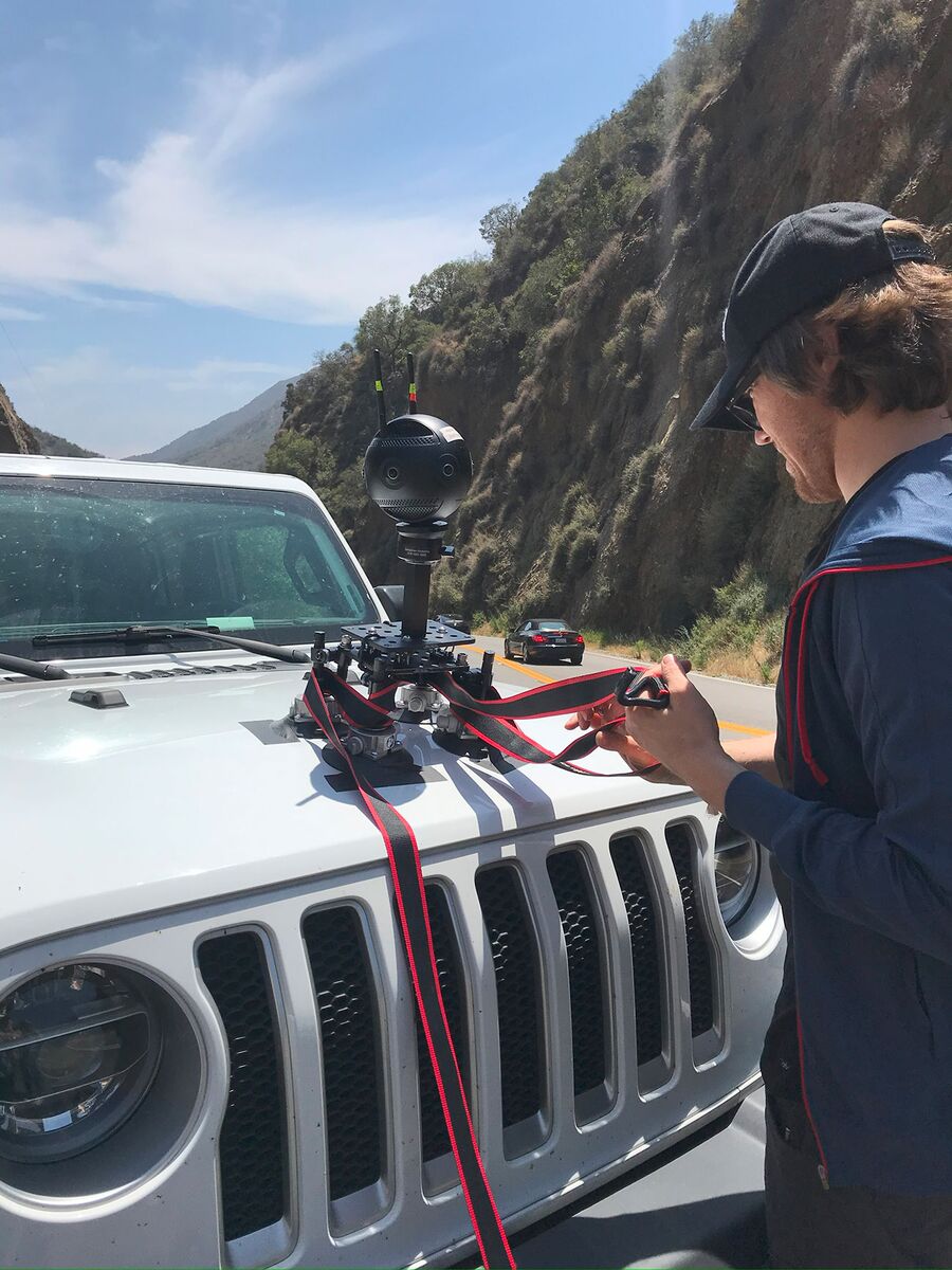 A crew member adjusts a 360 camera on a suction mount on the hood of a white Jeep on a canyon road