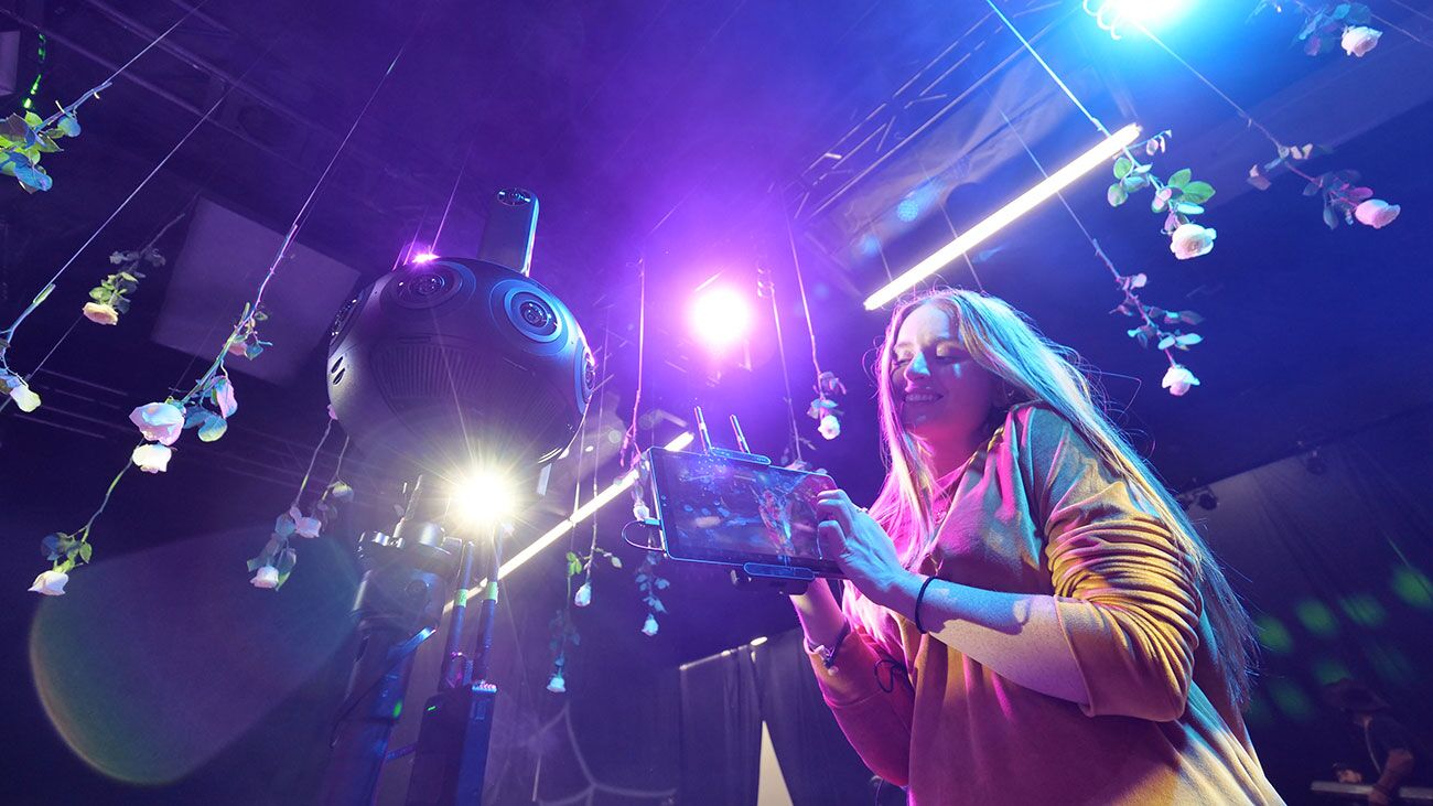 A dark music-video stage hung with white roses, a multi-lens 360 camera on a stand at left and a crew member checking a handheld monitor at right under purple light.