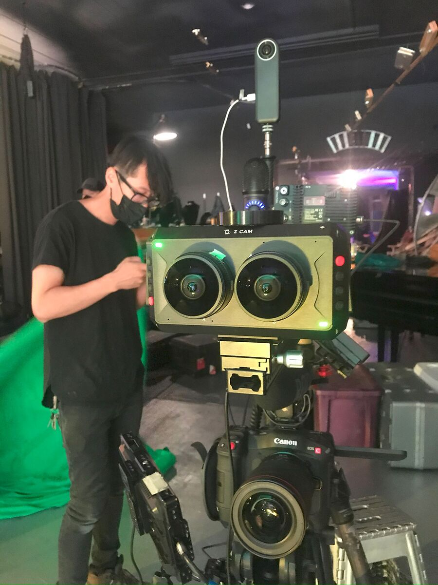 A crew member checks a dual-fisheye stereo camera stacked above a Canon cinema camera on a dark studio stage with a green screen