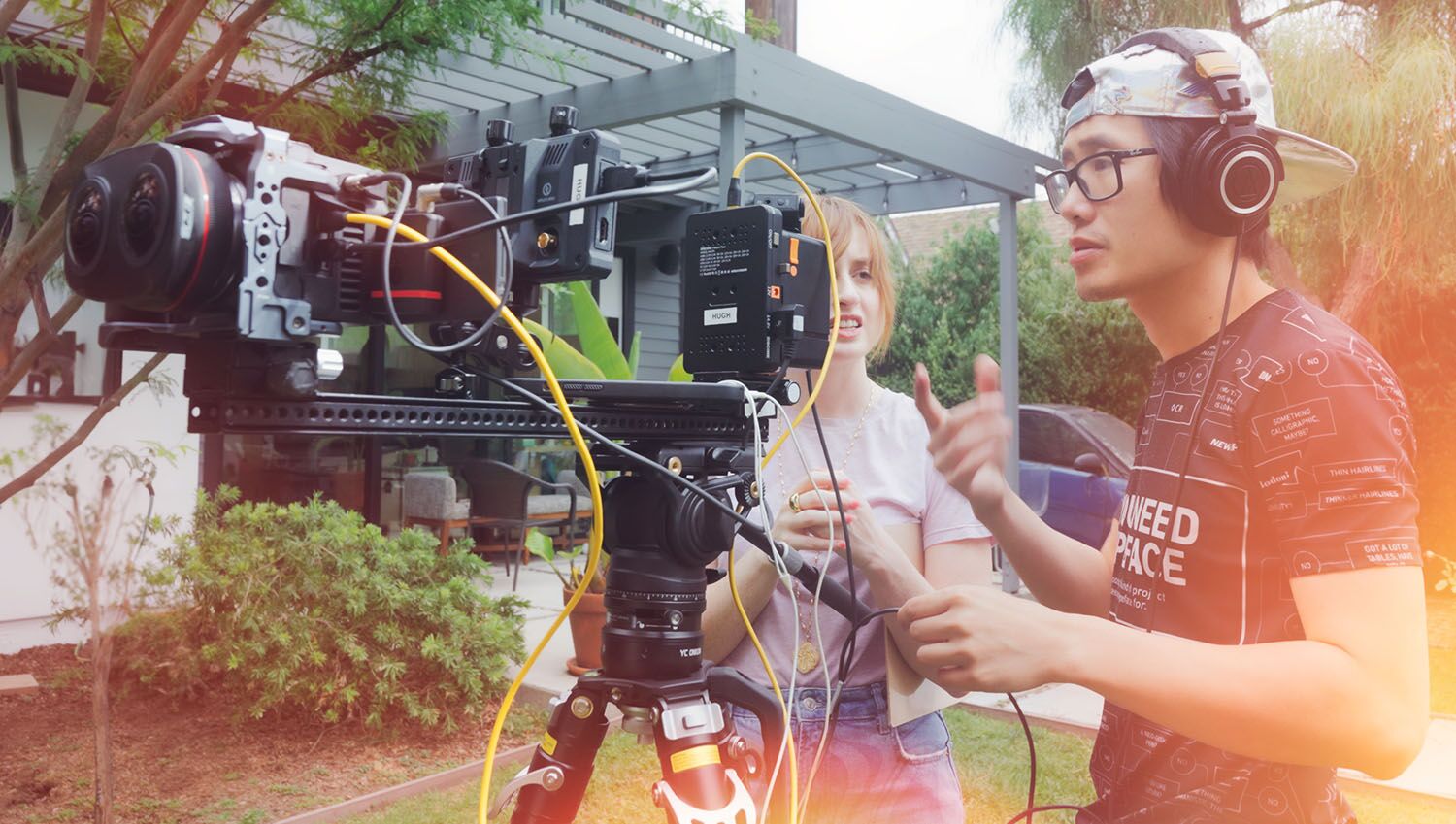 On set in a backyard: a woman and a man in a cap and glasses talk through a shot beside a stereo camera rig with a monitor and yellow cables.