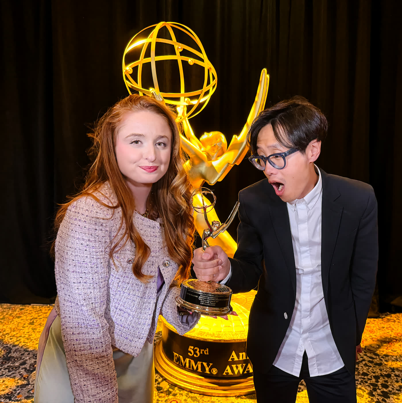 Keeley Turner and Hugh Hou holding an Emmy statuette in front of the giant gold Emmy statue at the 53rd Northern California Area Emmy Awards, Hugh pulling a surprised face.