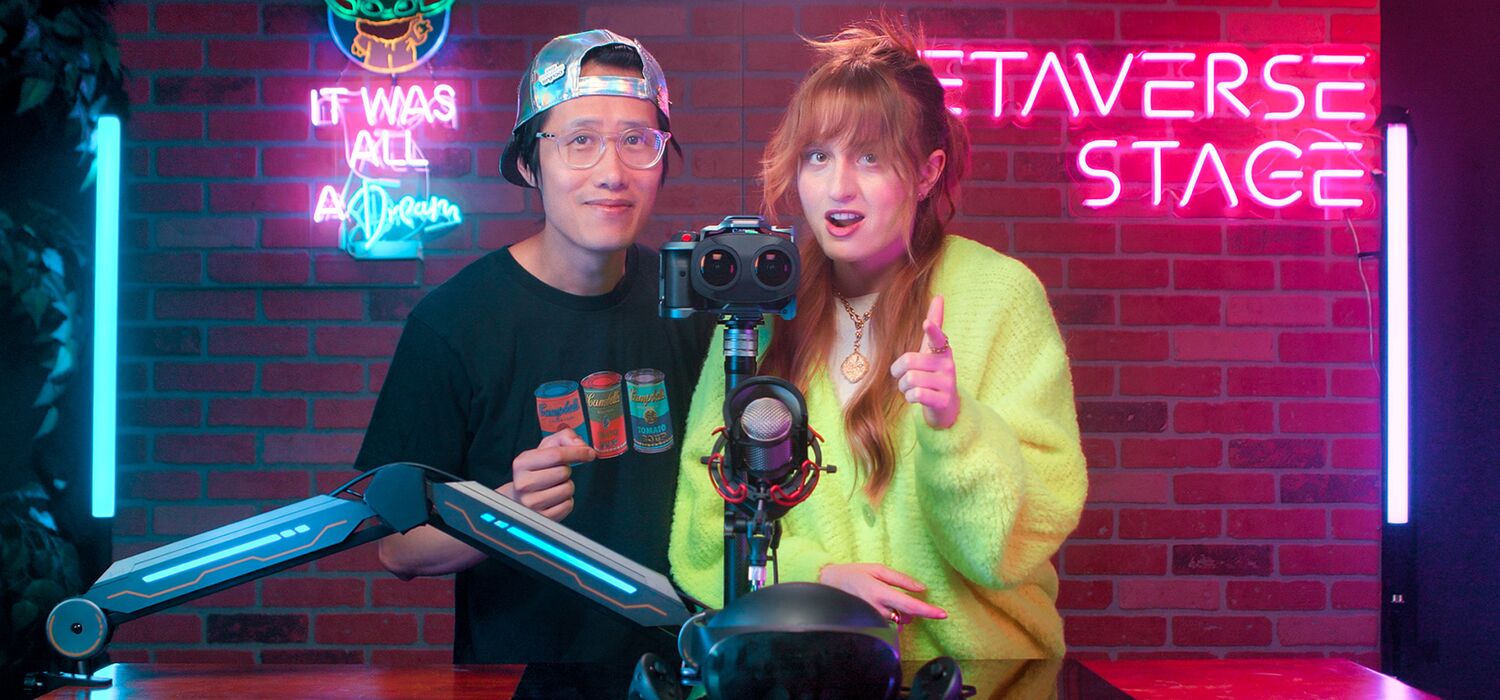 Hugh Hou and Keeley Turner behind a desk with a dual-lens 3D camera, under the studio's pink Metaverse Stage neon sign