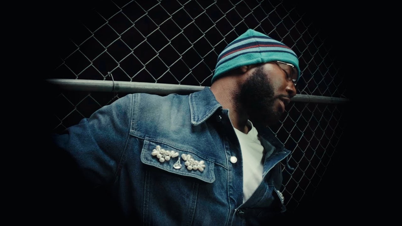 Khalid in a denim jacket and striped beanie leaning against a chain-link fence at night, from the Nah VR180 music video.